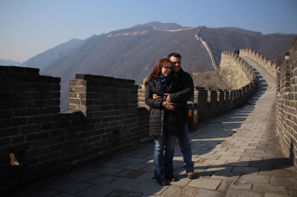 Alex and Bell are standing on a pathed section of the Great Wall of China with mountains and the wall trailing along them in the background