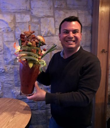 Alex holding Sobelmans Bloody Beast - a huge Bloody Mary pitcher garnished with a whole fried chicken, sliders, sausages and more!