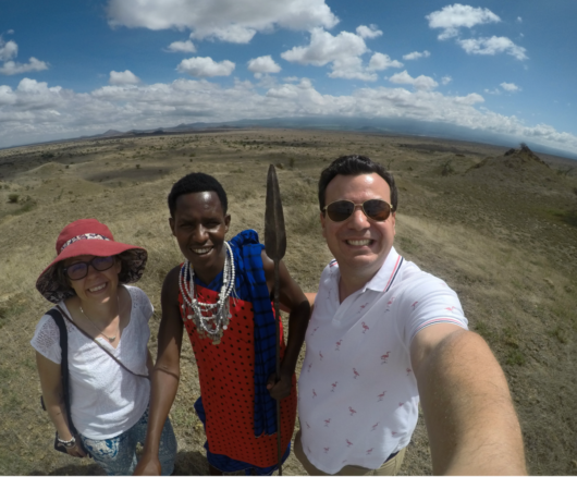 Selfie with a a Maasai warrior, and Alex and Bell, with dry plains and a a blue sky with white fluffy clouds