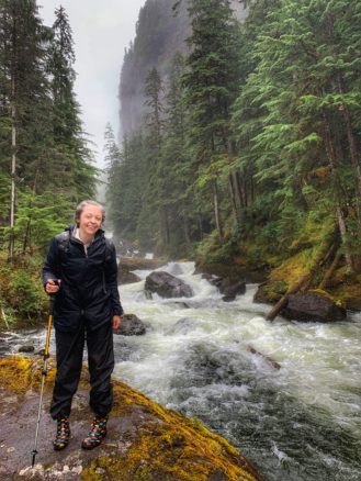 Bell in the rainforest in her rain gear with tall trees, misty skies and a fast moving river