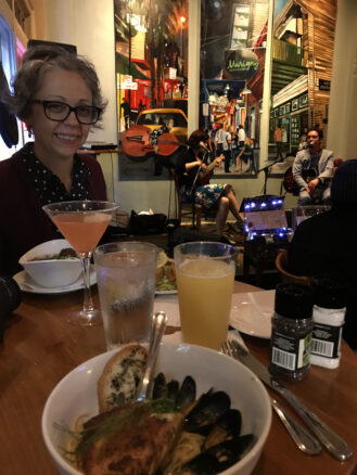 Bell at a table with drinks and mussels with art and musicians behind her