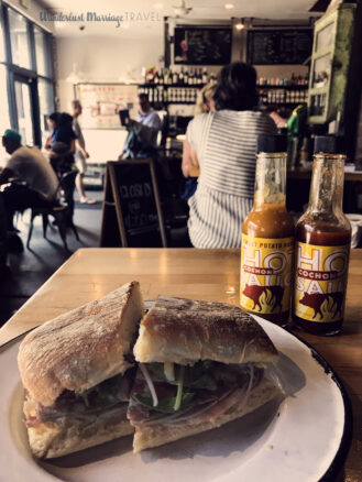 Muffaletta on a plate, with bottles of hot sauce and cafe in the background