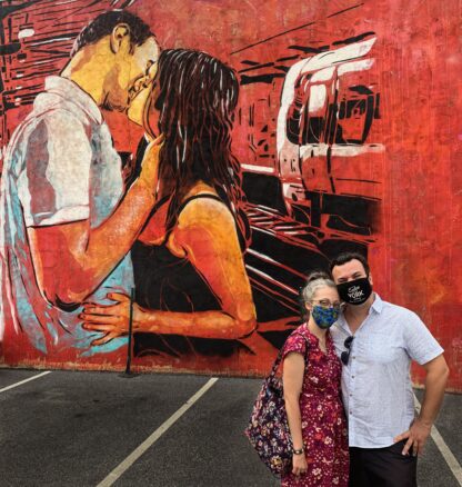 Alex & Bell with masks in front of a mural a guy kissing a girl 