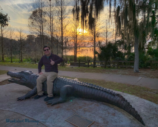 Alex sitting on a statue of a large alligator, with a lack in the background with the sun setting