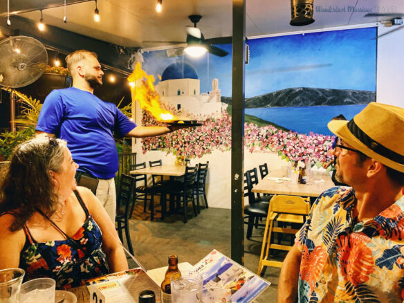 a man and a lady watch the restaurant server holding a plate with cheese on fire with a mural of Greece in the background