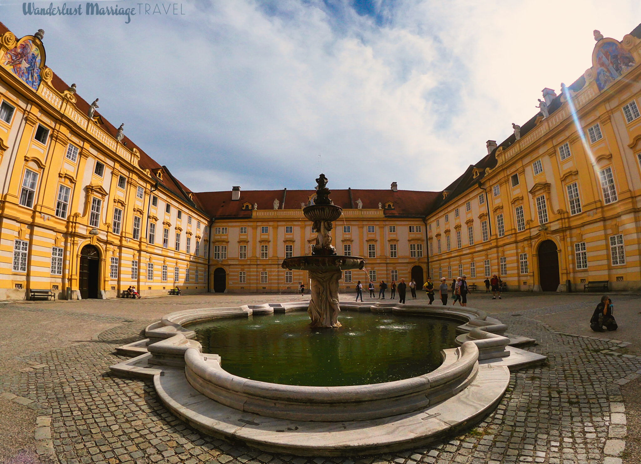 Photo of large courtyard with a fountain in the middle, the sun is shinning and people are walking around