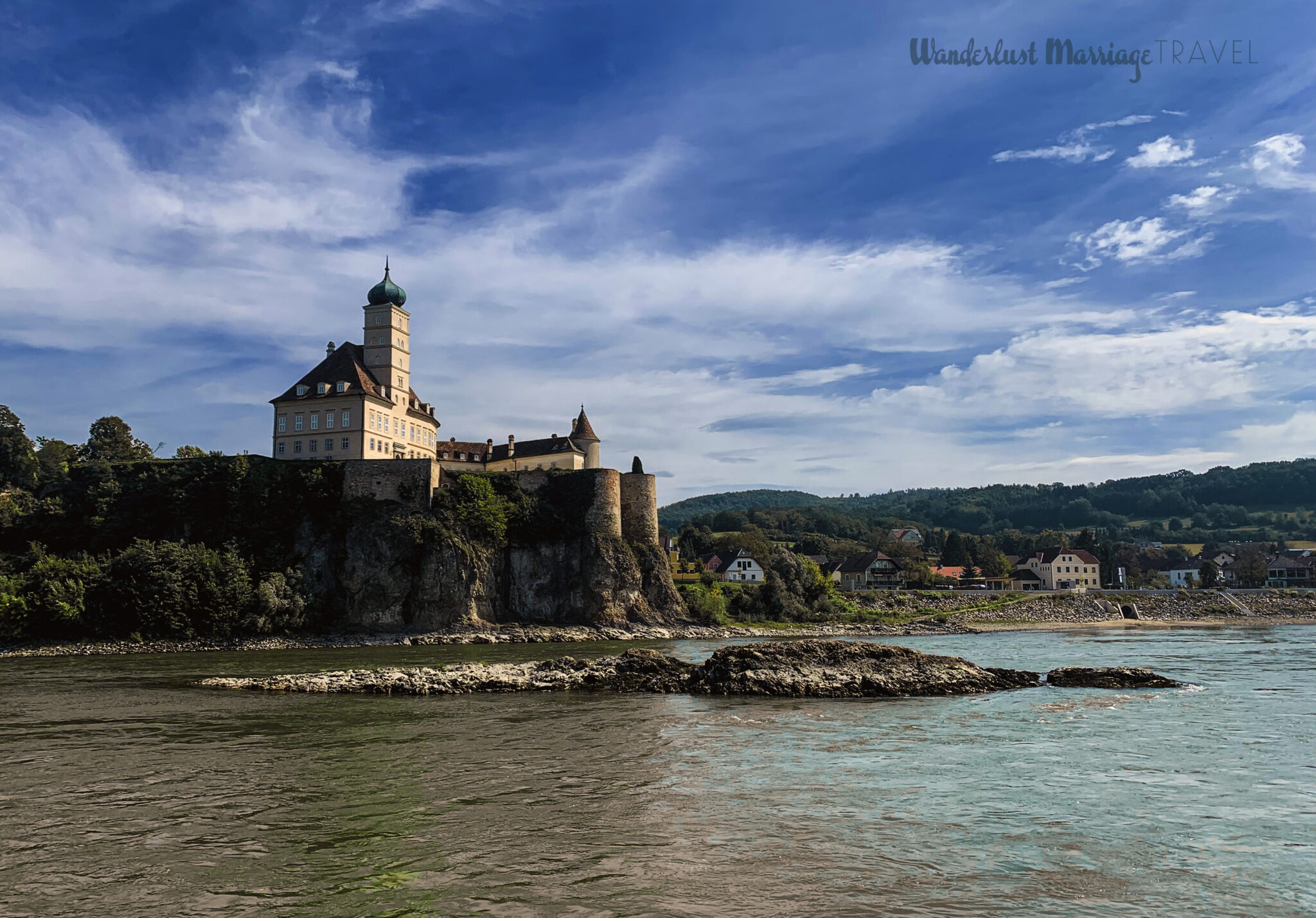 A castle has been built on the side of the river. It sits above the village. The sky is blue and there are hills in the background