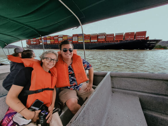 Alex and Bell are sitting in a small boat as a huge cargo ship sails behind them loaded up with shipping containers