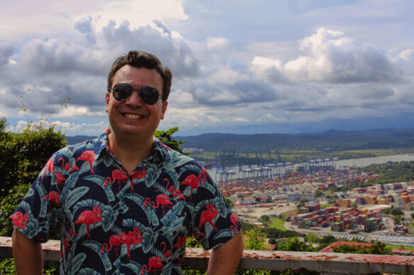 Alex in a Flamingo shirt is smiling with the cranes and shipping containers of the locks behind him, the sun in shinning