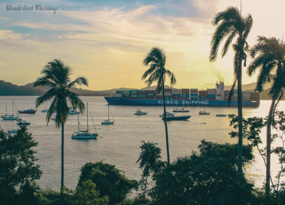 Large cargo ship coming out of the Panama Canal with little sail boats docked around and palm trees and the sun setting