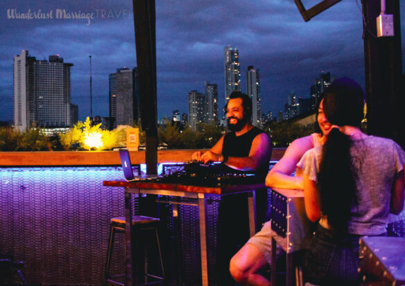 Man at a DJ stand smiling with the skyline of Panama lit up behind him, a couple are sitting at a table next to him