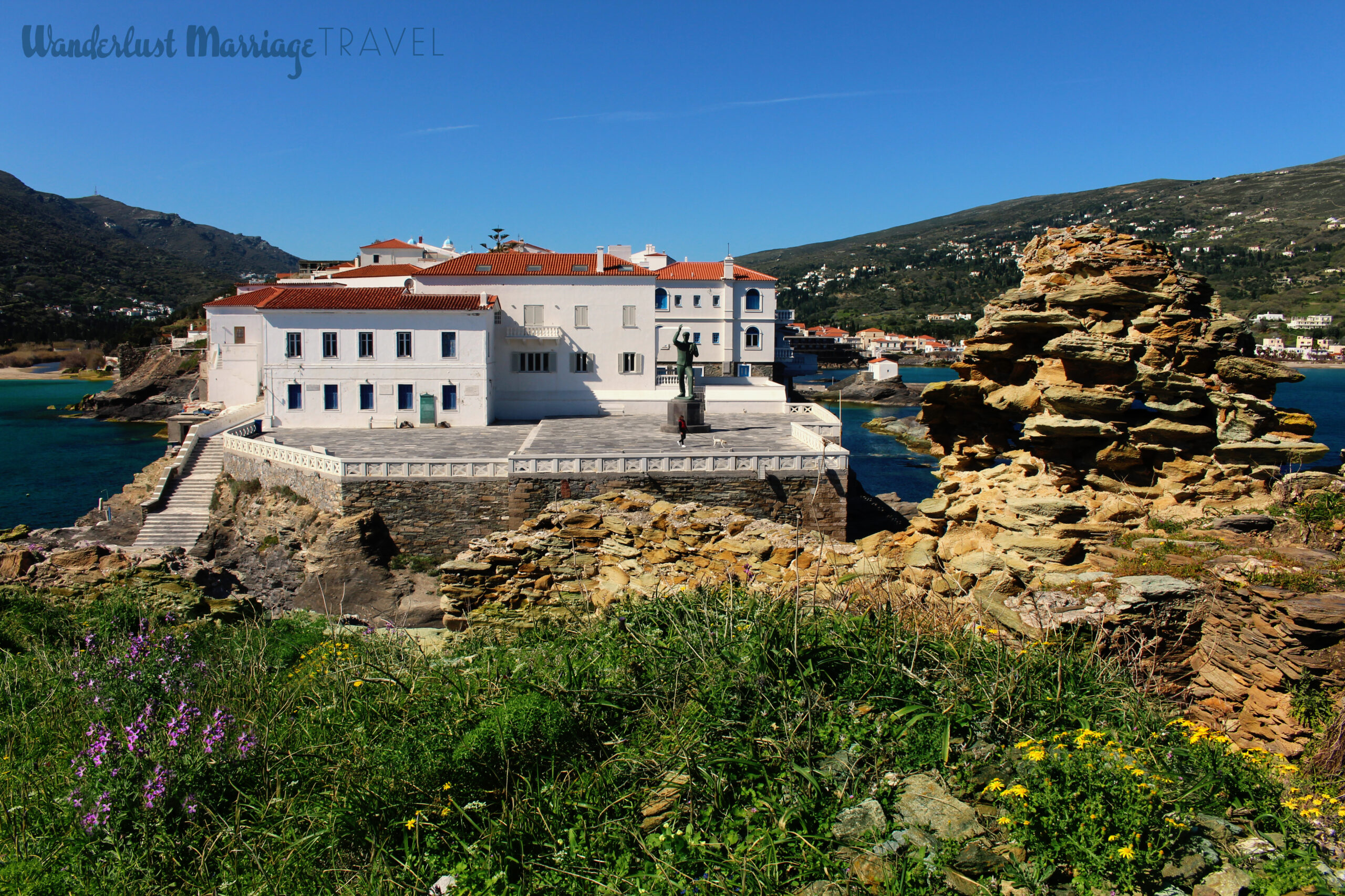 Ruins and wildflowers in the foreground, looking back on a village on a rock, surrounded by the sea and mountains