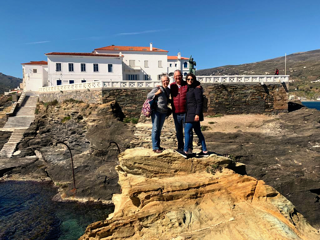 Two ladies and man stand on a rock with a white washed building in the background and the sea around the rock