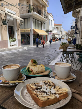 A table with bread with banana and coconut and a wrap and two coffees, with a pedestrian shopping street in the background