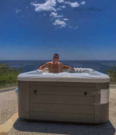 Alex sits in an above ground tub with the sea behind him and blue skies with a few wispy white clouds 