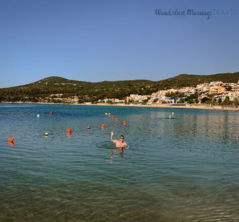 Alex is swimming in the sea and waving with the hills and village behind him