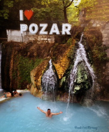 Alex is in a pool of light blue thermal water with a waterfall behind him with a sign saying I heart Pozar and the forest in the very background