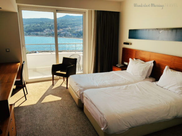 Hotel room with 2 twin bed, and a chair by the sliding door out to the balcony with a view of the sea and hills