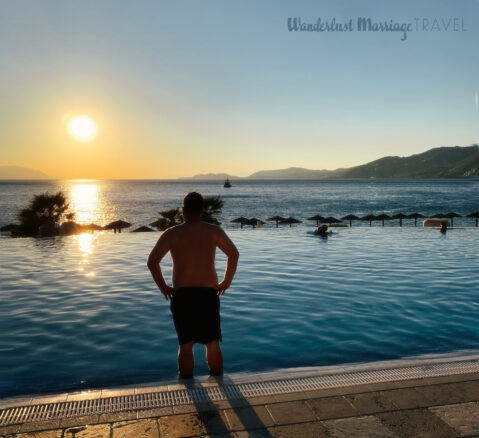 A man stands with his back to the camera with his feet in the infinity pool overlooking the sea and mountains as the sunsets