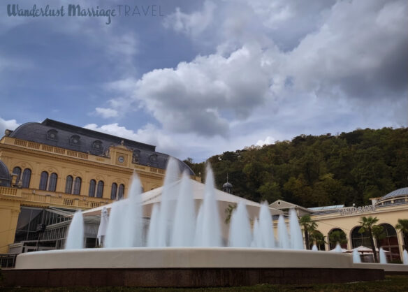 Fountain with a yellow building in the back grounds and a hill with many green trees, the sky is blue with some clouds