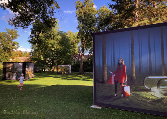 Large printed photo of girl coming up an escalator with shopping bags and her cell phone to find herself in a forest in  a park where a child plays 