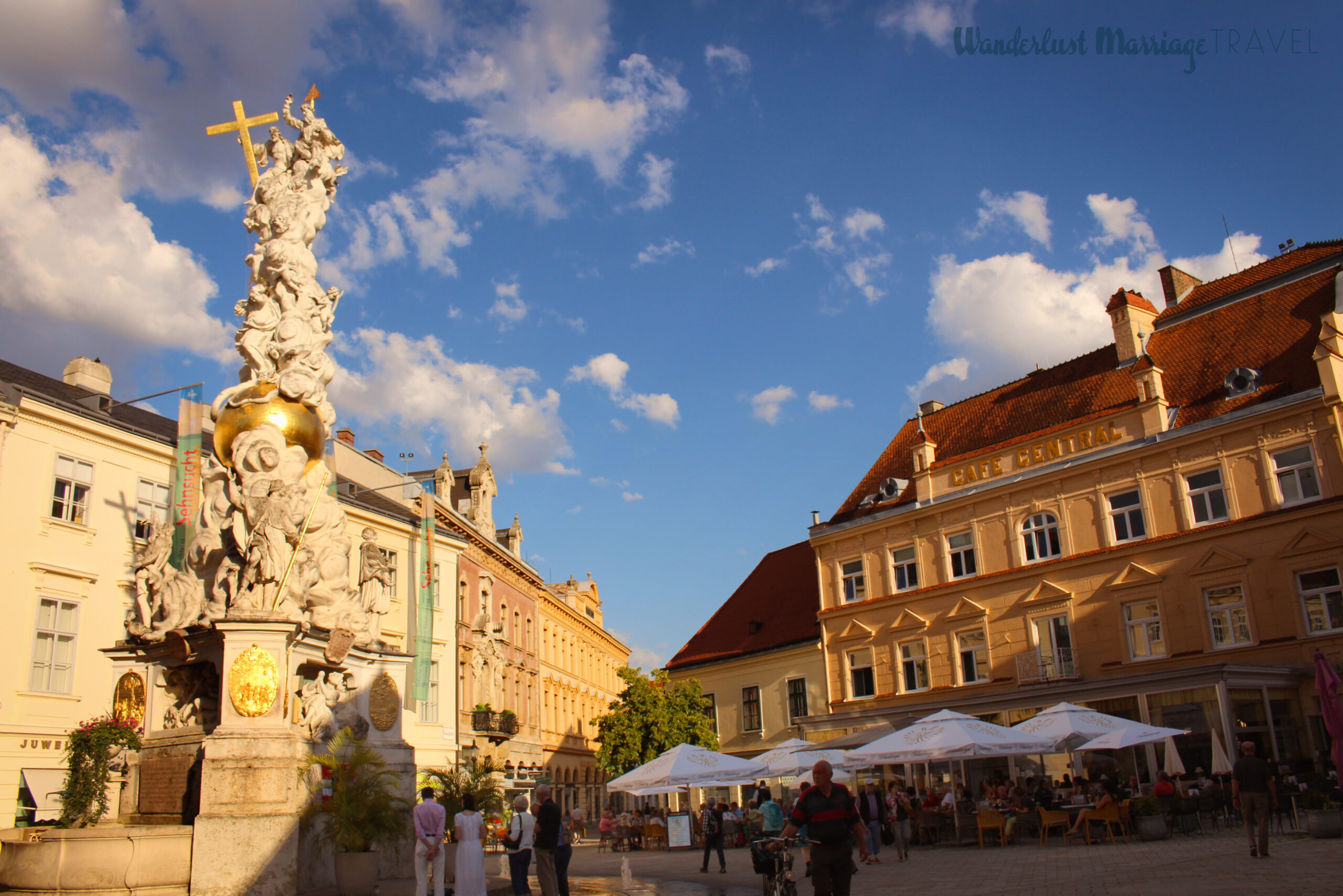 Statue of people a top a gold ball, a cafe with umbrella's out front and people walking around the square