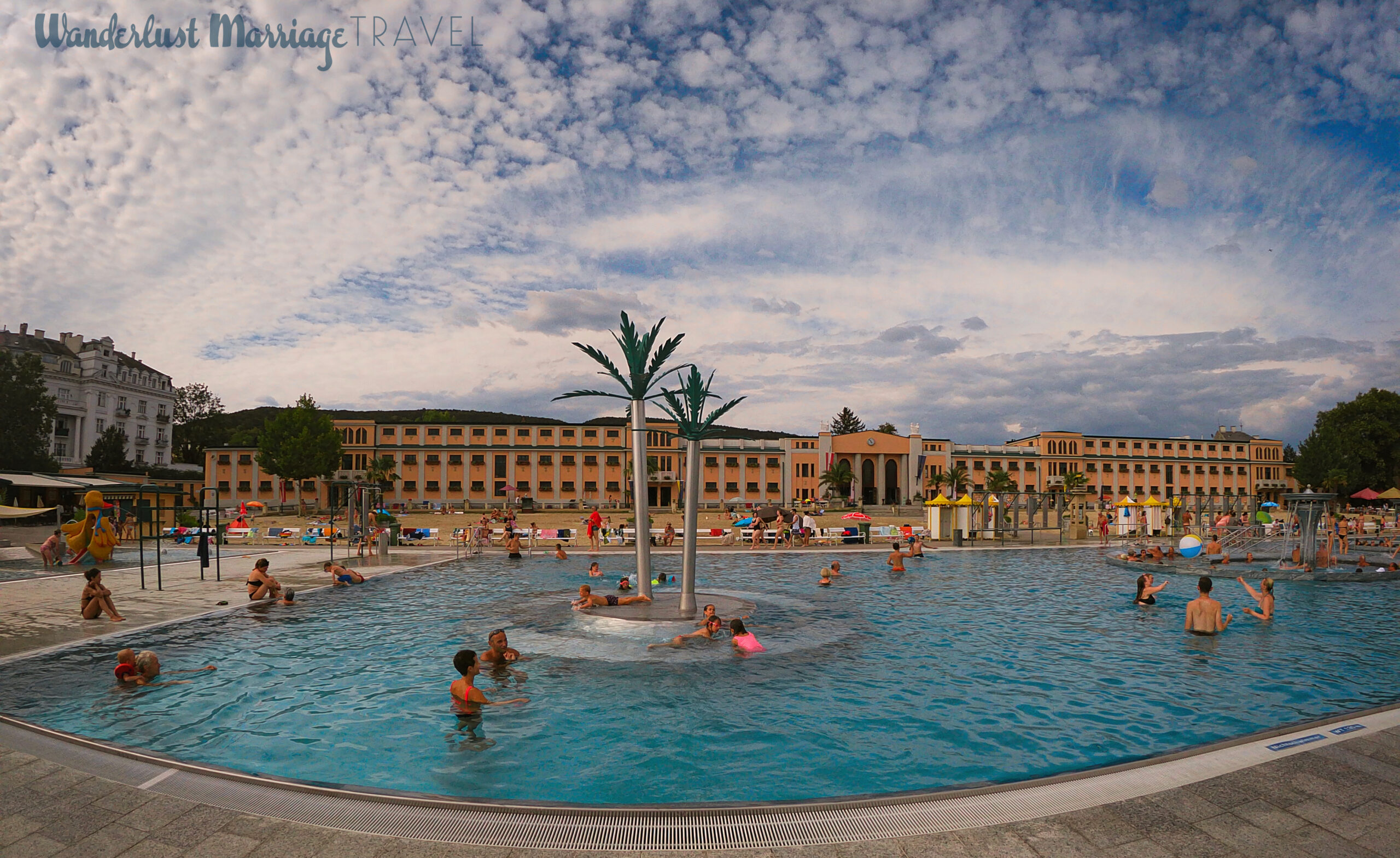 Large pool with metal palm trees in the middle, people swimming, beach in the background as well as an old style bath house