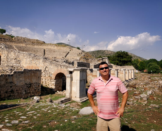 Alex stands in front of what remains of the amphitheater in Philipi with blue skies 