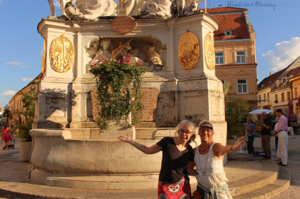 Two ladies stand in front of statue with their arms open, in the background people are walking around the town square