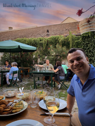 Alex sits at a table outside in a garden with a plate of Austrian meats and a glass of wine