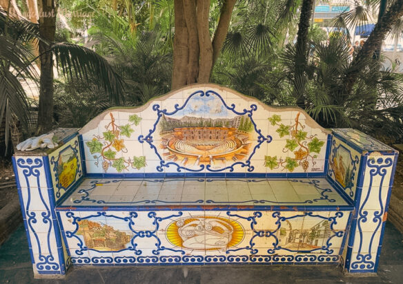 a bench that has colorful terracotta tiles in blue, white and yellow under some shady trees 