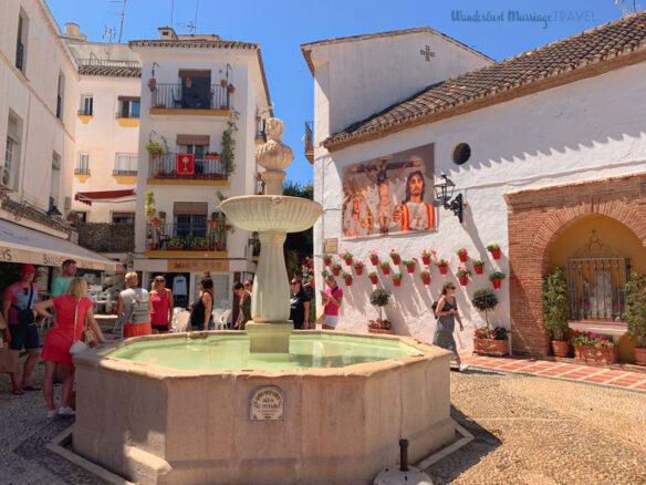 A water fountain in a small square surrounded by white-washed buildings and blue skies