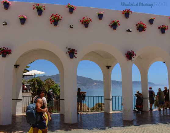 white washed walk way with pots of geranium flowers and the sea and mountains of the cost del sol in the background