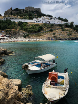 Fortress a top a hill overlooking the bay in Lindos, with 2 boats floating in the crystal clear waters