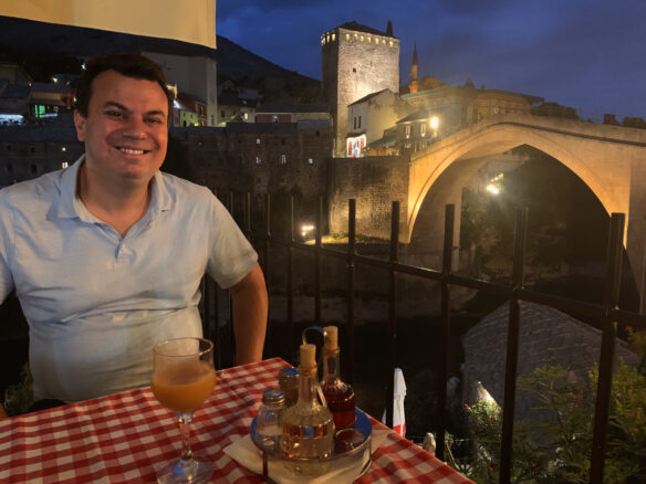 Alex sits at a dinner table with a red and white checker table cloth with the Stari Most bridge in the background