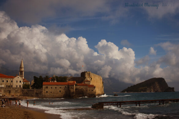Landscape shot of the beach with people swimming in the sea and a pier, with a small island of in the background as well as old city walls of Budva