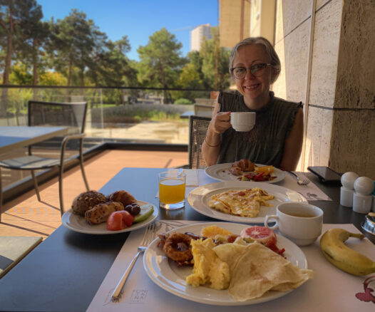 Bell is at the breakfast table with coffee, eggs, pastries and fresh fruit with beautiful blue skies behind her