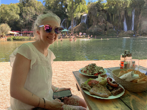 Bell sits at a table with a chicken salad and meat platter with waterfalls and people swimming behind her