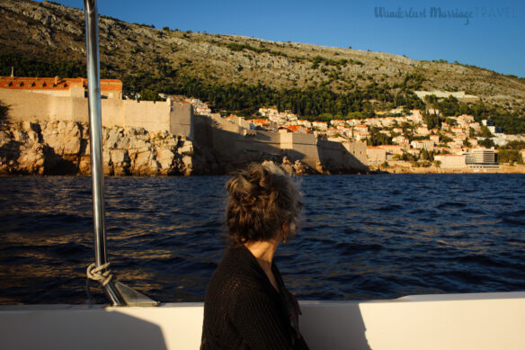 Lady looks out over the blue sea at the stone wall surrounding the old town, with a mountain and blue skies in the background