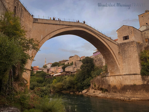 High arched bridge built from stone crossing a river with the village in the background