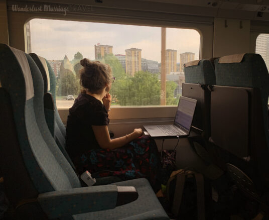 Bell is sitting in a train carriage with her laptop working remotely with the city passing by out the window