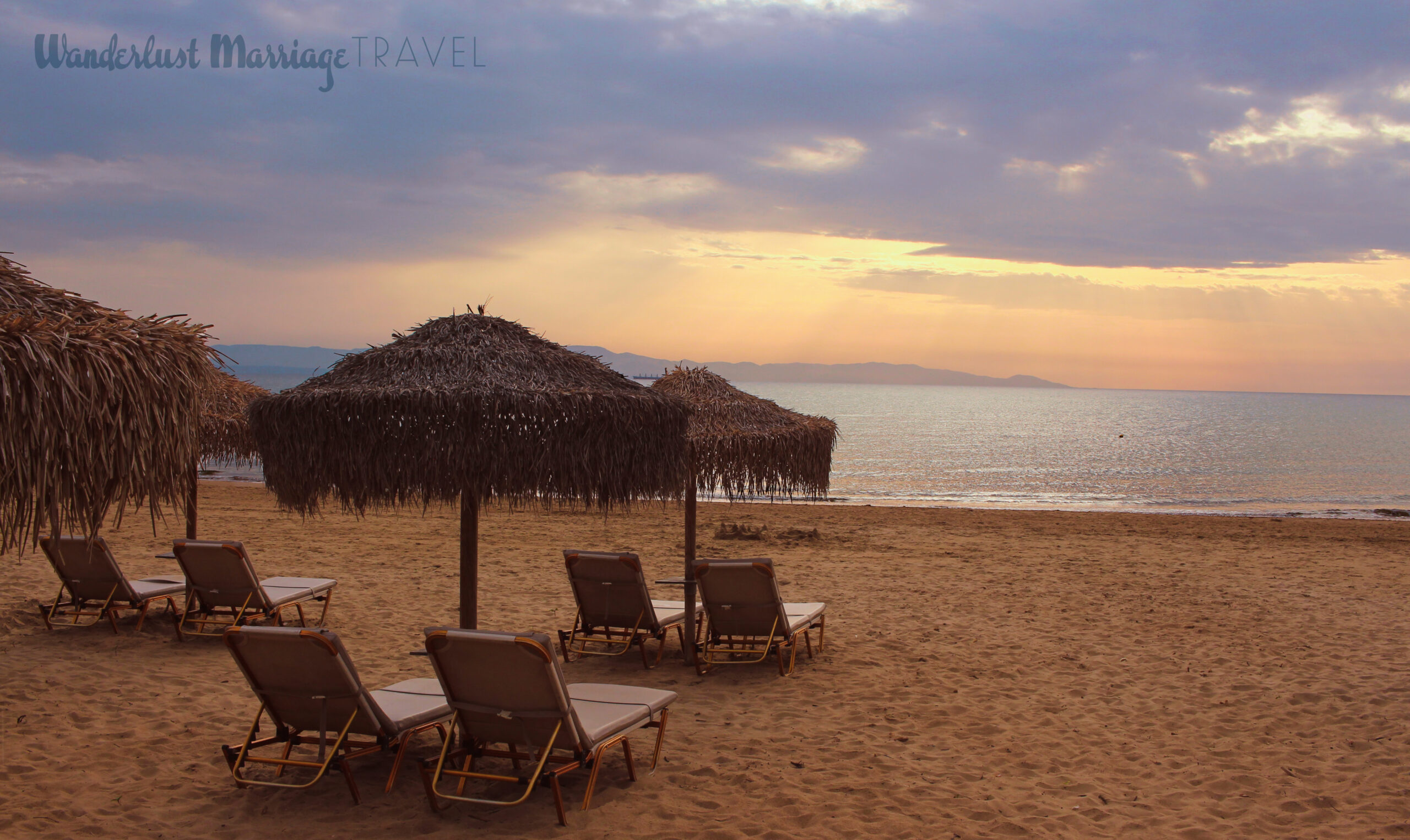 Sunset on the beach with lounge chairs and rustic grass umbrellas that are part of the Greco Olympia Resort