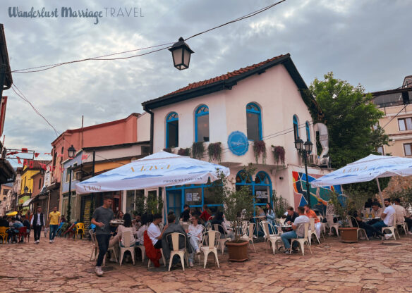 Cobble stone streets intersecting with a cafe on the corner and people sitting and drinking coffee
