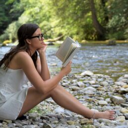 An attractive young brunette girl reading a book barefoot on rocks next to a stream in the woods.