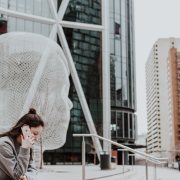 A young lady talking on her phone in the middle of a city, in front of a modern art installation.