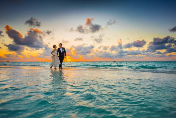 A bride and groom walk along the water on the beach with a colorful sunset in the background.