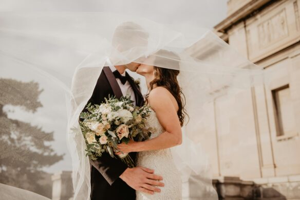 A couple kissing after taking their wedding vows, with their faces blocked by a veil. The bride is holding a lovely bouquet of flowers.