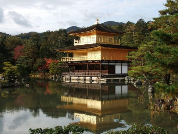 The Golden Pavilion of Kinkaku-ji Temple in Kyoto, Japan reflects off the lake, with many trees behind it.