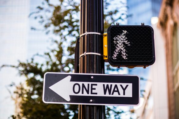 A digital pedestrian cross sign with a white person indicating it's safe to walk, above a one way sign.