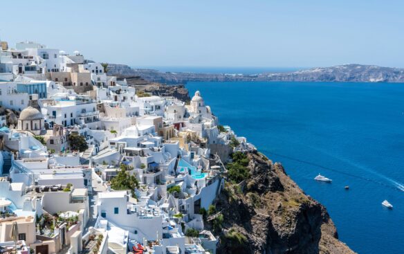 Predominantly blue and white buildings dot the cliffs of Santorini, Greece nestle alongside the blue waters of the Aegean Sea.